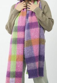 Personne portant un pull beige et tenant une écharpe douce à carreaux multicolores rose, violet, vert et orange, suspendue devant.