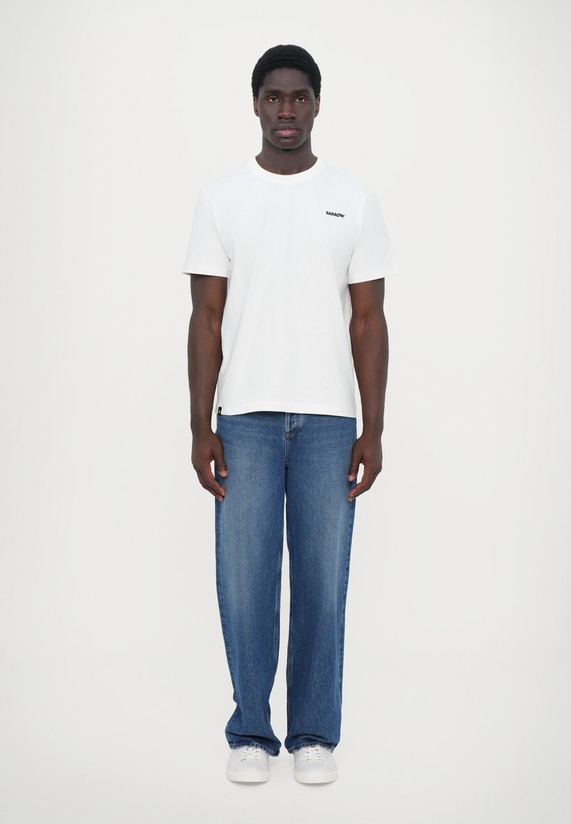 T-shirt en coton blanc avec un logo noir, associée à un jean en denim bleu ample et des baskets blanches. Design simple, sans motifs ou accents distincts.
