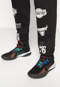 Svarta sportskor med flerfärgade snören, överdel i mesh och texturerad sula. Svarta träningsbyxor med olika sportlogotyper i vitt.