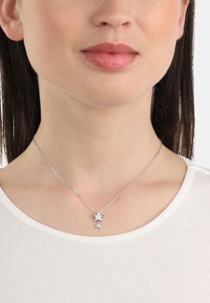 Collar de plata con dos colgantes en forma de estrella de diferentes tamaños; cadena delicada con una textura suave; llevado sobre una blusa blanca de canalé.