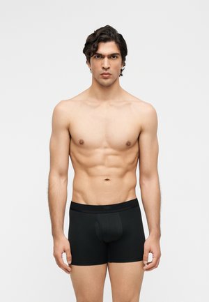 Jeune homme en forme aux cheveux bouclés foncés, debout face à l’avant, portant un boxer noir sur un fond clair uni.