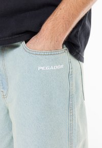 Világoskék farmer rövidnadrág kőmosott textúrával; fehér "PEGADOR" márkázással, öt zsebes dizájnnal és kontrasztos varrással rendelkezik.