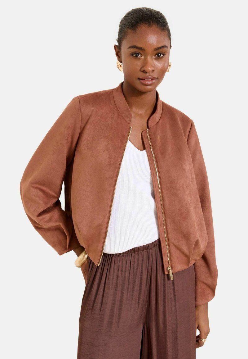 Žena v hnedej semišovej bunde so zipsom, bielom rebrovanom tielku a voľných hnedých nohaviciach na jednoduchom svetlom pozadí.