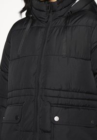 Musta toppatakki, jossa korkea kaulus, vetoketjusuljenta ja kaksi etutaskua, joissa nepparit. Pinnan kuviointi vaakasuuntaisilla tikkauksilla.