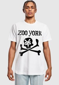 Bílý bavlněný tričko s potiskem "ZOO YORK" v černé barvě nad grafikou lebky a zkřížených kostí, s volným střihem a kulatým výstřihem.