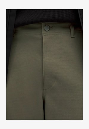 Pantaloni verde oliva in tessuto con una leggera trama a quadri, dotati di chiusura con bottone davanti e passanti per cintura, abbinati a un top nero strutturato.