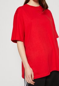 Červené oversized tričko vyrobené z bavlny, s krátkými rukávy, kulatým výstřihem a jemným designem tří pruhů na rukávech.