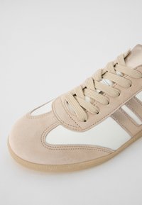 Sneaker con tomaia in suede beige, dettagli in pelle bianca, lacci tonali e suola in gomma chiara. Tre strisce orizzontali sui lati.