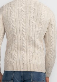 Homme portant un pull crème en maille torsadée et un jean bleu, vu de dos sur un fond uni clair.