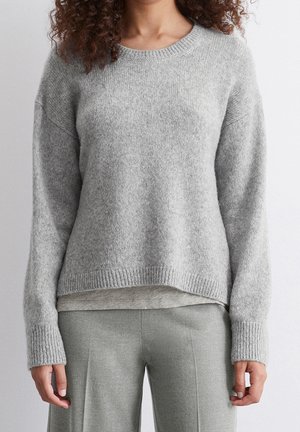 Persoon draagt een lichtgrijze gebreide trui over een licht overhemd, gecombineerd met grijze op maat gemaakte broek tegen een effen witte achtergrond.