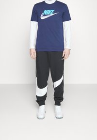 Námořnické tričko Nike s světle modrým logem, vrstvené přes bílé tričko s dlouhým rukávem; černé teplákové kalhoty s bílými zakřivenými detaily; bílé obuv.