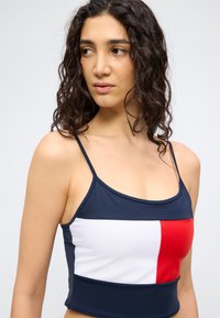 Νεαρή γυναίκα με σγουρά σκούρα μαλλιά που φοράει τοπ σε σκούρο μπλε με λευκά και κόκκινα ορθογώνια χρωματικά μπλοκ, κοιτάζοντας προς το πλάι.