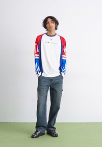 Hvid langærmet skjorte med rød og blå flamme-design, logo på brystet, parret med blå jeans og sorte sko. Ensfarvet baggrund.