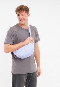 Világoskék válltáska kerek formával, puha szövettextúrával és logópacával. Keresztben hordva.