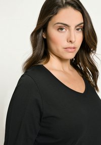 Černý dlouhý rukáv s výstřihem do V vyrobený z měkké tkaniny, s uvolněným střihem a mírně texturovaným povrchem. Obličej modelu je zobrazen, zaměřen na detaily.