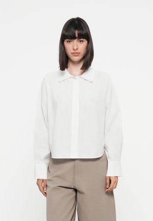 Weißes Baumwollhemd mit Knopfleiste, spitzen Kragen, langen Ärmeln und einem cropped Design. Kombiniert mit beigen Schlaghosen.
