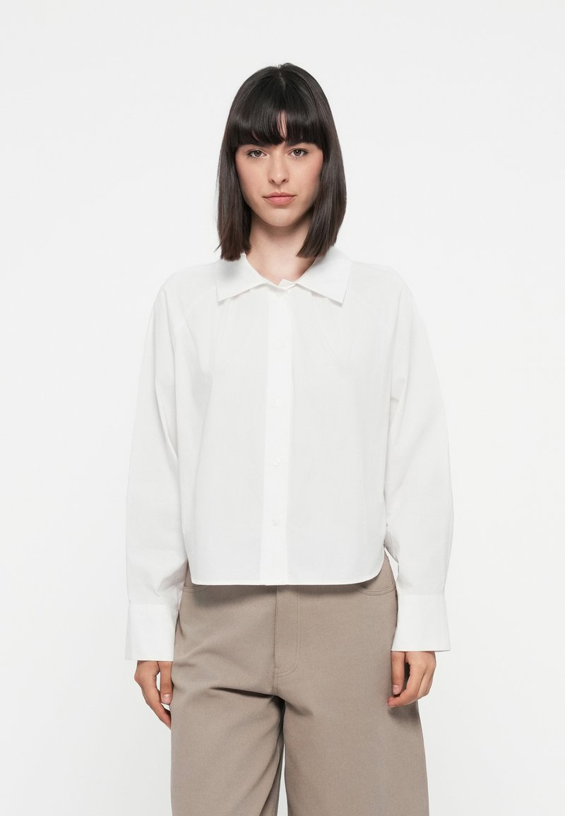 Camicia bianca in cotone con colletto a punta, maniche lunghe e design cropped. Abbinata a pantaloni beige a gamba larga.