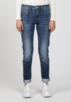 Person wearing cuffed blue jeans with faded front and white sneakers, standing against a plain light background.