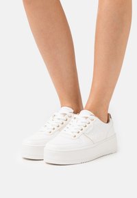 Weiße Plateausneaker mit strukturierten Akzenten, goldenen Ösen und einer glatten Oberfläche. Mit einer robusten Sohle und klassischem Schnürdesign.