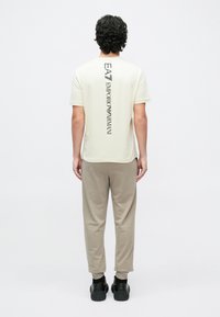 Egy férfi háttal áll, krémszínű rövid ujjú pólót visel, amelyen függőlegesen olvasható az "EA7 Emporio Armani" logó, bézs színű melegítőnadrágot és fekete sportcipőt visel.