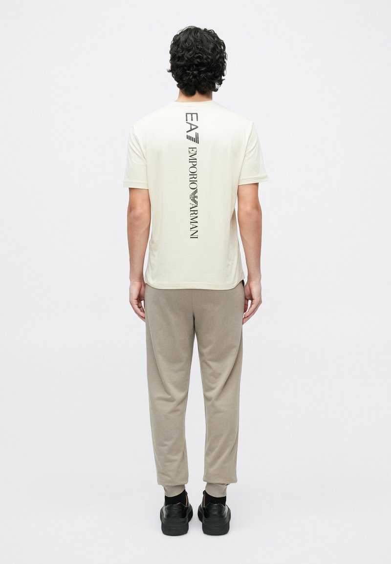 Egy férfi háttal áll, krémszínű rövid ujjú pólót visel, amelyen függőlegesen olvasható az "EA7 Emporio Armani" logó, bézs színű melegítőnadrágot és fekete sportcipőt visel.