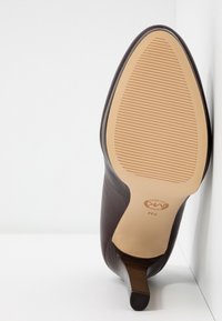 Chaussure à talon haut en cuir marron inclinée vers le haut, montrant une semelle beige avec de fines lignes horizontales antidérapantes et la pointe du talon sur une surface blanche.