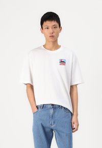 White cotton t-shirt with short sleeves, round neckline, and a square graphic patch featuring blue and red colors on the chest.