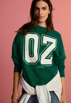 Sweatshirt vert avec un grand imprimé blanc "02" et le texte "Wicked" en vert, avec des poignets et une taille côtelés, superposé sur un tissu gris.