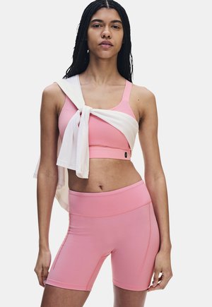 Vrouw draagt een roze sportbeha en bijpassende bikershorts met een witte trui gestrikt rond haar schouders tegen een effen achtergrond.