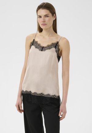 Kvinde iført en beige silkeundertrøje med sort blonderkant og sorte bukser, stående mod en ensartet lys baggrund.