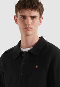 Polo shirt lavorato a maglia nero con colletto, caratterizzato da un logo ricamato arancione e dettagli dei bottoni, ambientato su uno sfondo grigio chiaro.
