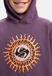 Lila hoodie med ett stort grafiskt motiv av en orange sol med svarta detaljer och en central logotyp. Materialet verkar mjukt med en avslappnad passform.