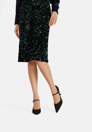 Scarpe nere in suede con tacco alto, punta affusolata e cinturini alla caviglia, abbinate a una gonna al ginocchio verde e nera scintillante con paillettes.