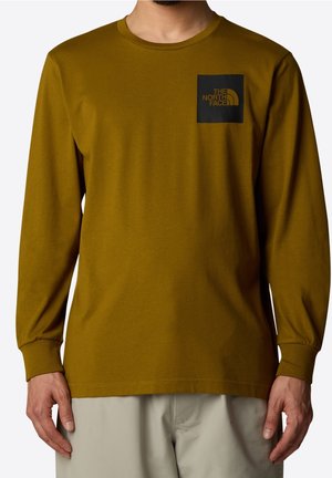 Mężczyzna w musztardowożółtej koszuli z długim rękawem, z czarnym kwadratowym logo i napisem "The North Face" na lewej piersi.