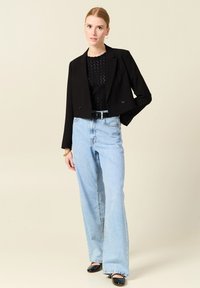 Schwarze kurze Blazerjacke mit doppelten Knöpfen, kombiniert mit hellblauen, hochgeschnittenen, weit geschnittenen Jeans. Das Outfit wird durch schwarze Schuhe ergänzt.