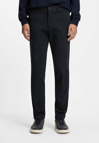 Czarne spodnie o dopasowanym kroju, z płaskim przodem i dwoma bocznymi kieszeniami. Noszone z ciemnymi sneakersami, mają gładkie wykończenie i subtelną fakturę.