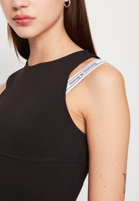 Svart ärmlös topp med rundad halsringning och vita märkesstrappar med "Tommy Jeans." Slät tyg med en figurnära design.