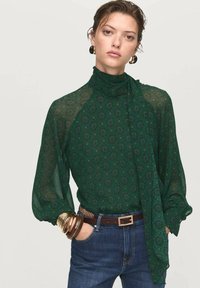 Blusa verde con patrón, de cuello alto y mangas largas abullonadas, combinada con jeans oscuros y pulseras doradas. Tejido texturizado con una calidad semitranslúcida.