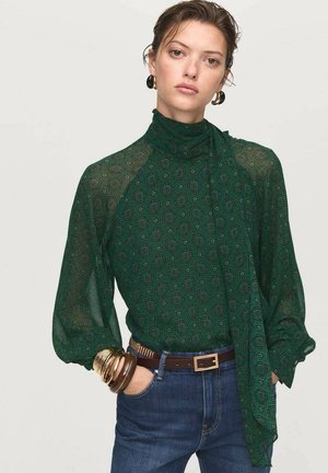 Blouse verte à motifs avec un col montant et des manches bouffantes longues, associée à un jean sombre et des bracelets en or. Tissu texturé avec une qualité semi-transparente.