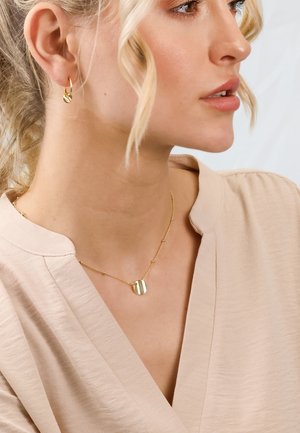 Goldene Halskette mit einem runden Anhänger und vertikalen Streifen, getragen mit einem hellbeigen Oberteil. Kleine goldene Creolen vervollständigen den Look.