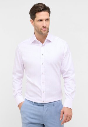 Mann mit braunen Haaren, der ein hellrosa Hemd zum Knöpfen und hellblaue Hose trägt, stehend mit einer Hand in der Tasche.