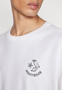 Prim-plan al gâtului și părții superioare a pieptului purtând un tricou alb cu logo-ul negru Converse, care prezintă o stea și două mâini ridicate.