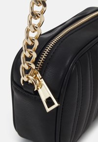 Borsa in pelle nera con finitura testurizzata, tracolla in catena dorata e dettagli della cerniera in vista. Forma rettangolare compatta con accenti trapuntati.