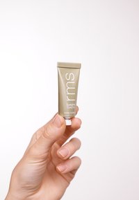 Tube doré de produit pour les yeux étiqueté "rms Eyelights," avec du texte blanc. Main tenant le tube contre un fond blanc.