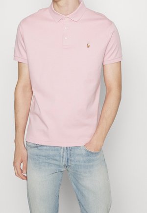 Man draagt een lichtroze korte mouwen poloshirt met een klein geborduurd logo op de borst en lichtblauwe spijkerbroek.