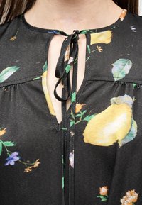 Μαύρη μπλούζα με κίτρινο μοτίβο λεμονιών και πράσινα φύλλα, με λαιμόκοψη σε σχήμα κλειδώματος δεμένη με λεπτό υφασμάτινο φιόγκο.