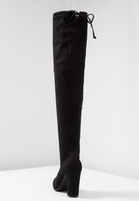 Bota de piel de ante negra, de muslo alto, con tacón grueso y lazada en la parte superior, de pie sobre una superficie blanca contra un fondo gris claro.