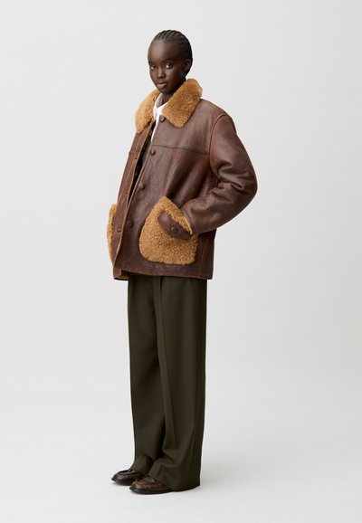 Chaqueta de piel marrón con cuello de piel sintética color beige y bolsillos. Corte holgado con cierre de botón. Un par de pantalones verde oliva debajo.