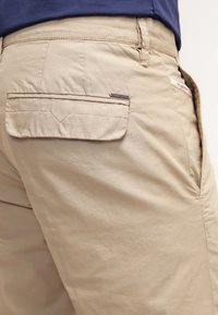 Pantalons beiges avec une poche arrière à rabat et une petite étiquette de marque en métal sur la poche, portés avec une chemise navy.
