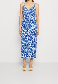 Donna che indossa un tutù senza maniche di colore bianco con stampa floreale blu e cravatta in vita, abbinato a sandali bianchi con cinturini intrecciati.